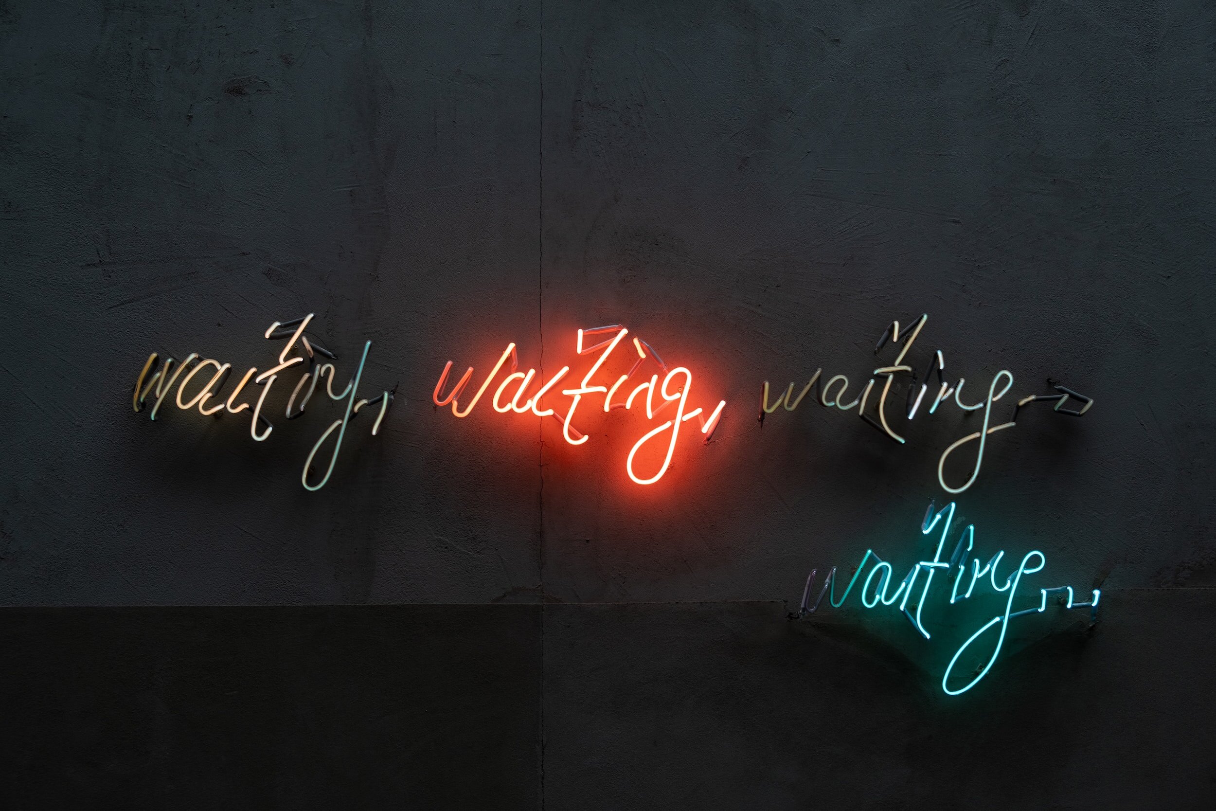 Four neon signs on a dark wall spell out the word “waiting” in different colors and sizes.