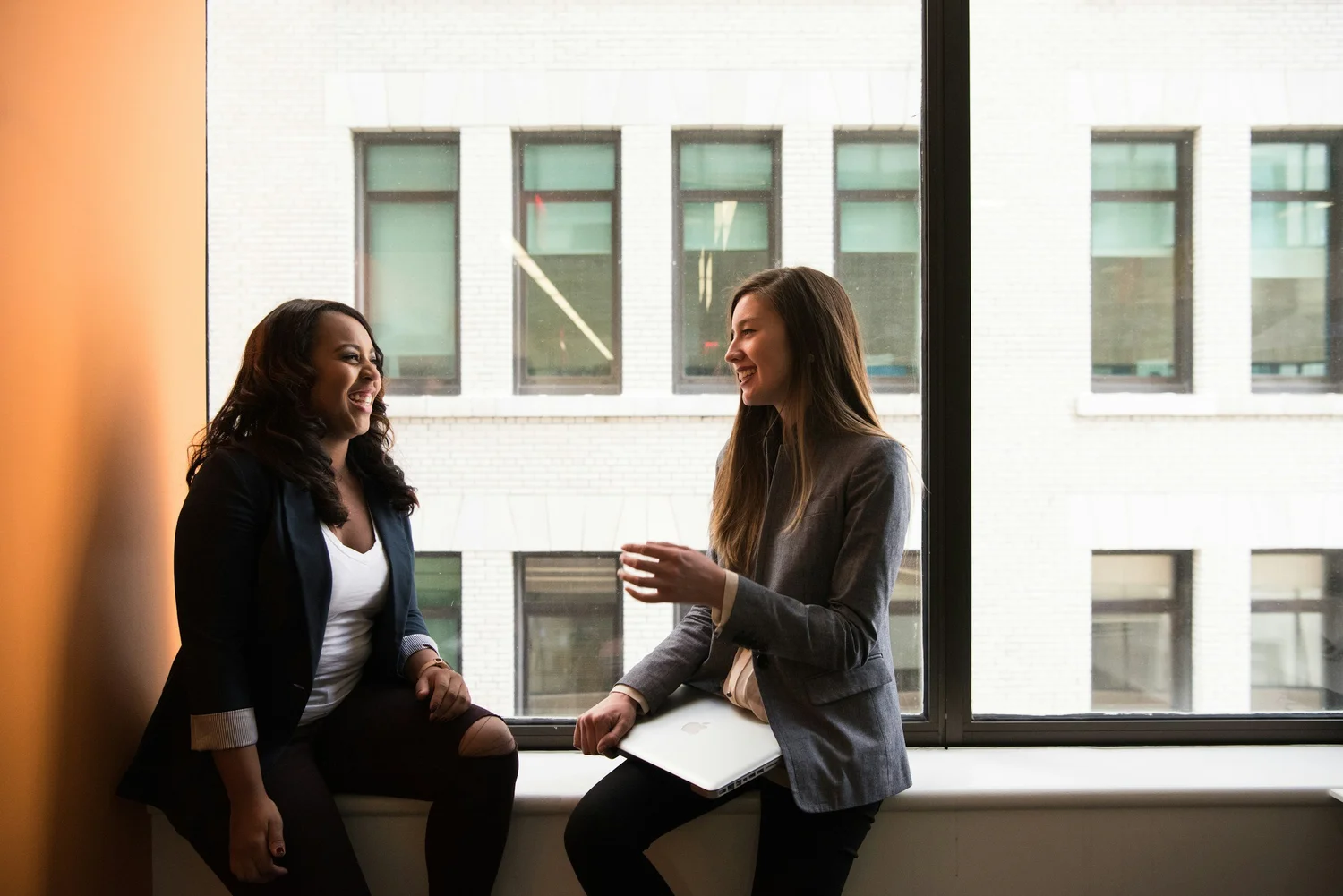 Two women in business attire laughing while sitting by a window with a laptop.