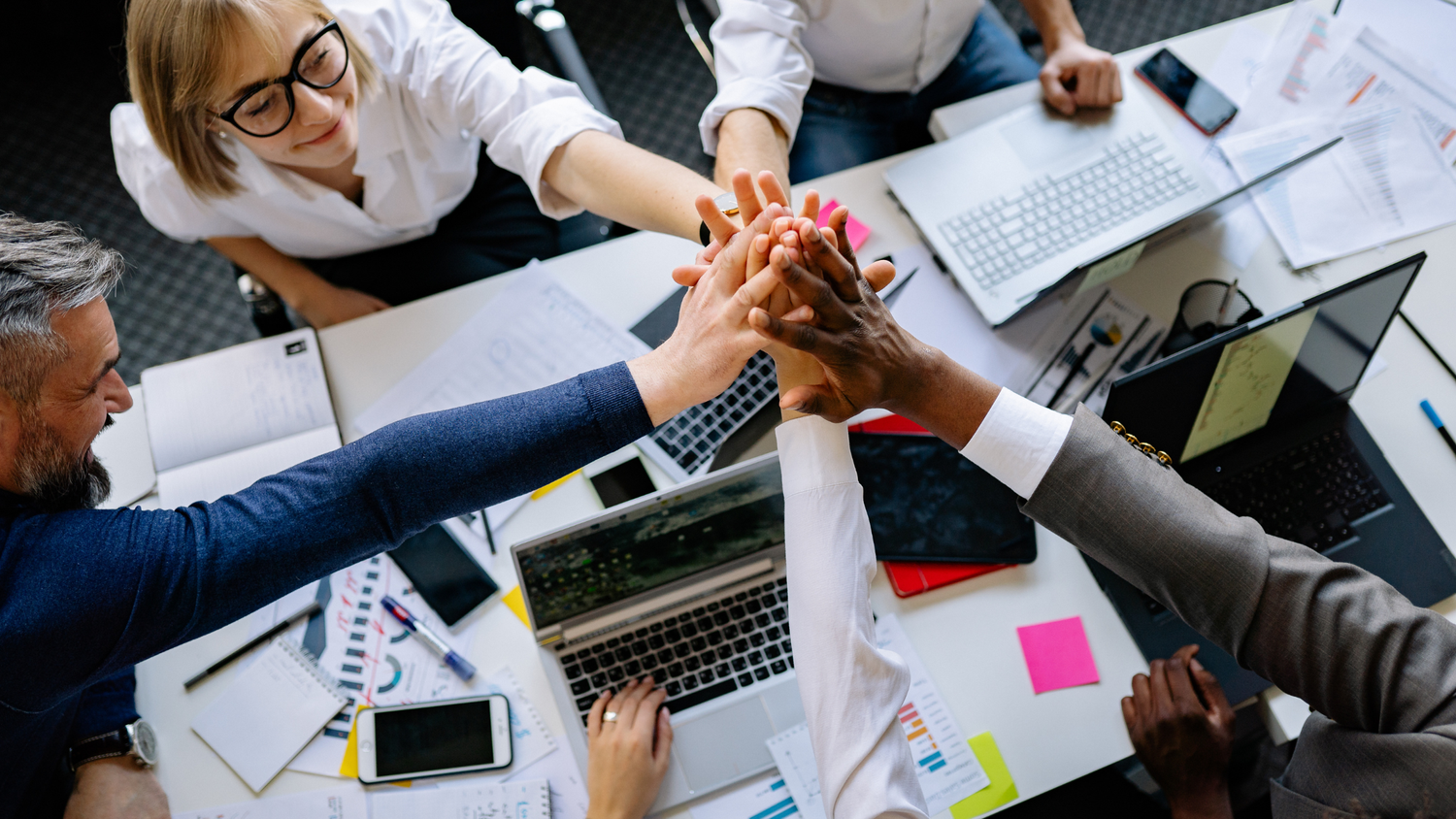 A group of diverse professionals at a table with laptops and papers put their hands together in a sign of unity.