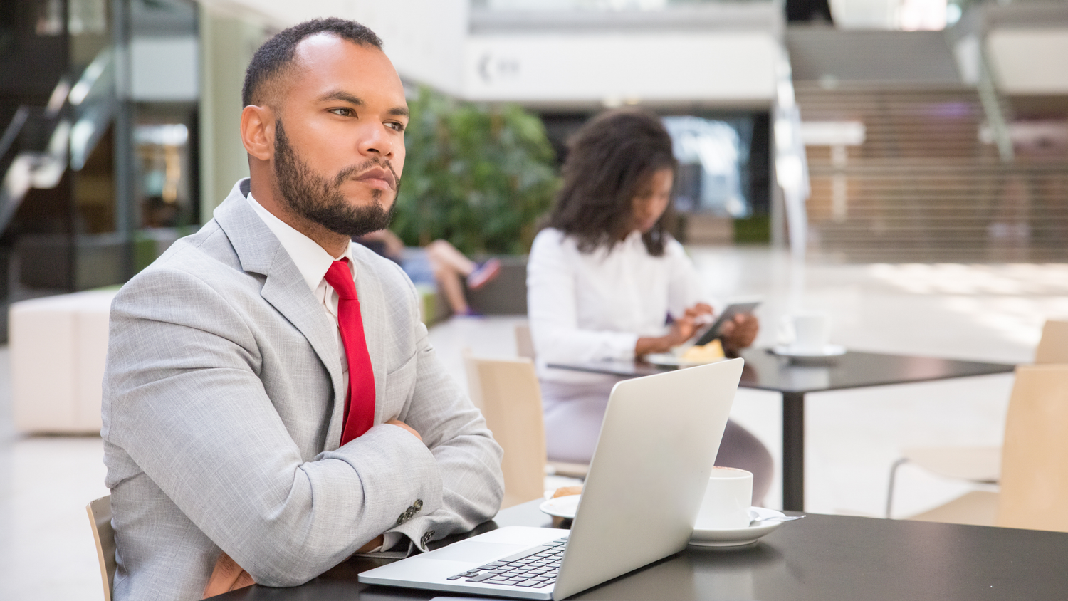Man in a gray suit and red tie with arms crossed sits at a table with a laptop; a woman in the background looks at a tablet.