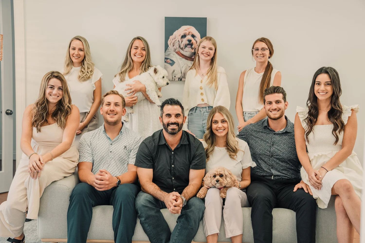 A team of 10 people and 2 dogs pose for a photo on a couch, with a painting of a dog in an astronaut suit on the wall.