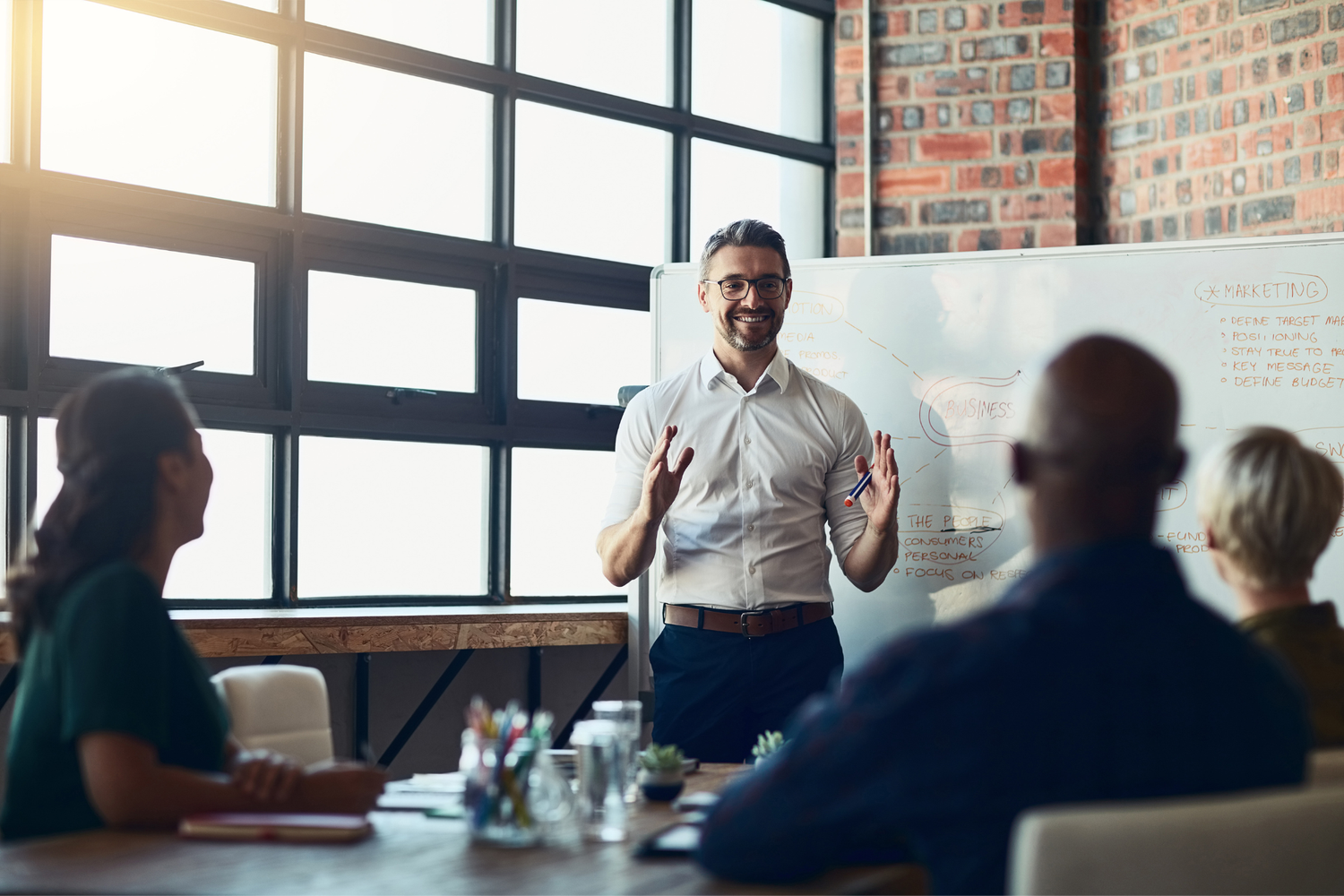 Smiling presenter at a whiteboard discussing marketing strategy with colleagues in a meeting room.
