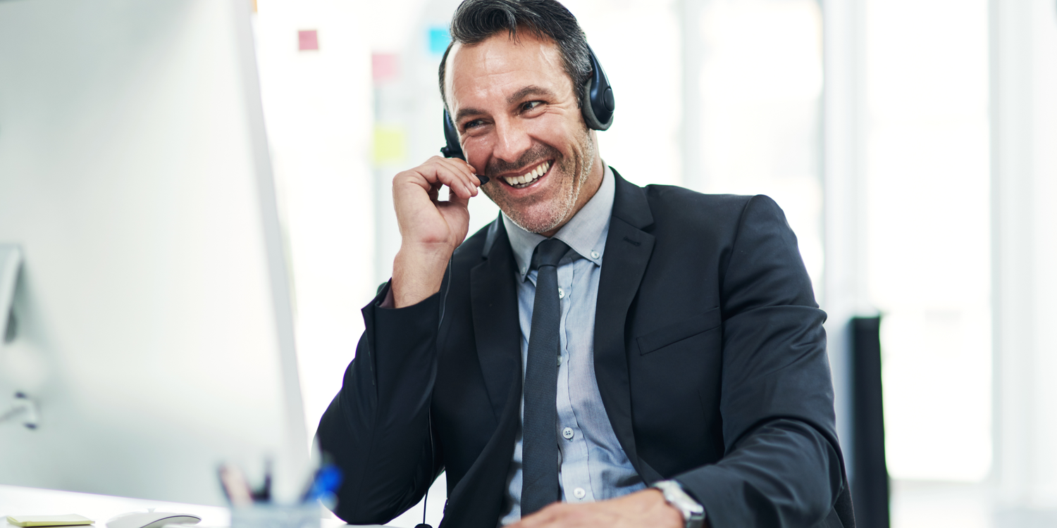 A smiling man in a suit wearing a headset answers the phone at his desk.