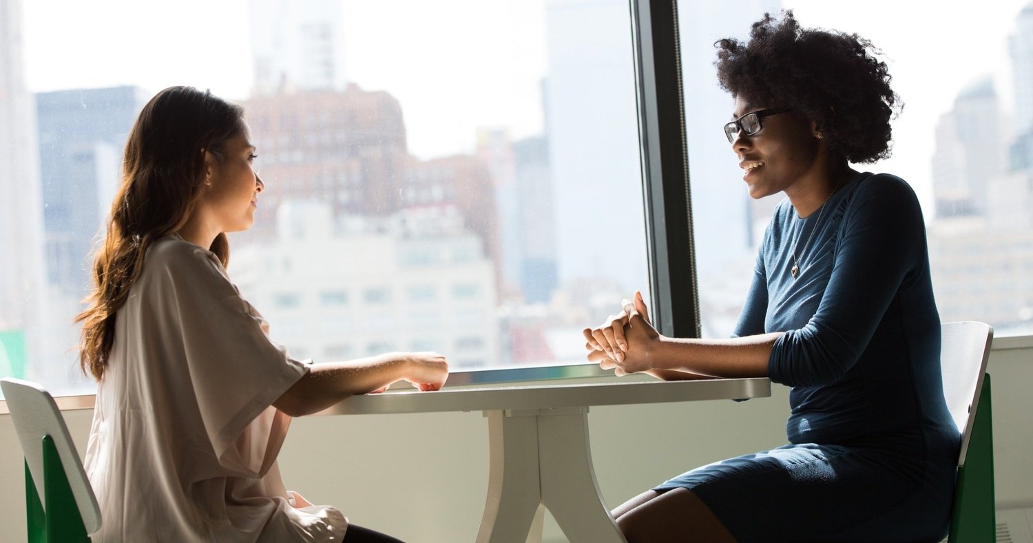 Two women sit at a table talking; one has long dark hair in a blush top, the other has dark curly hair and glasses.