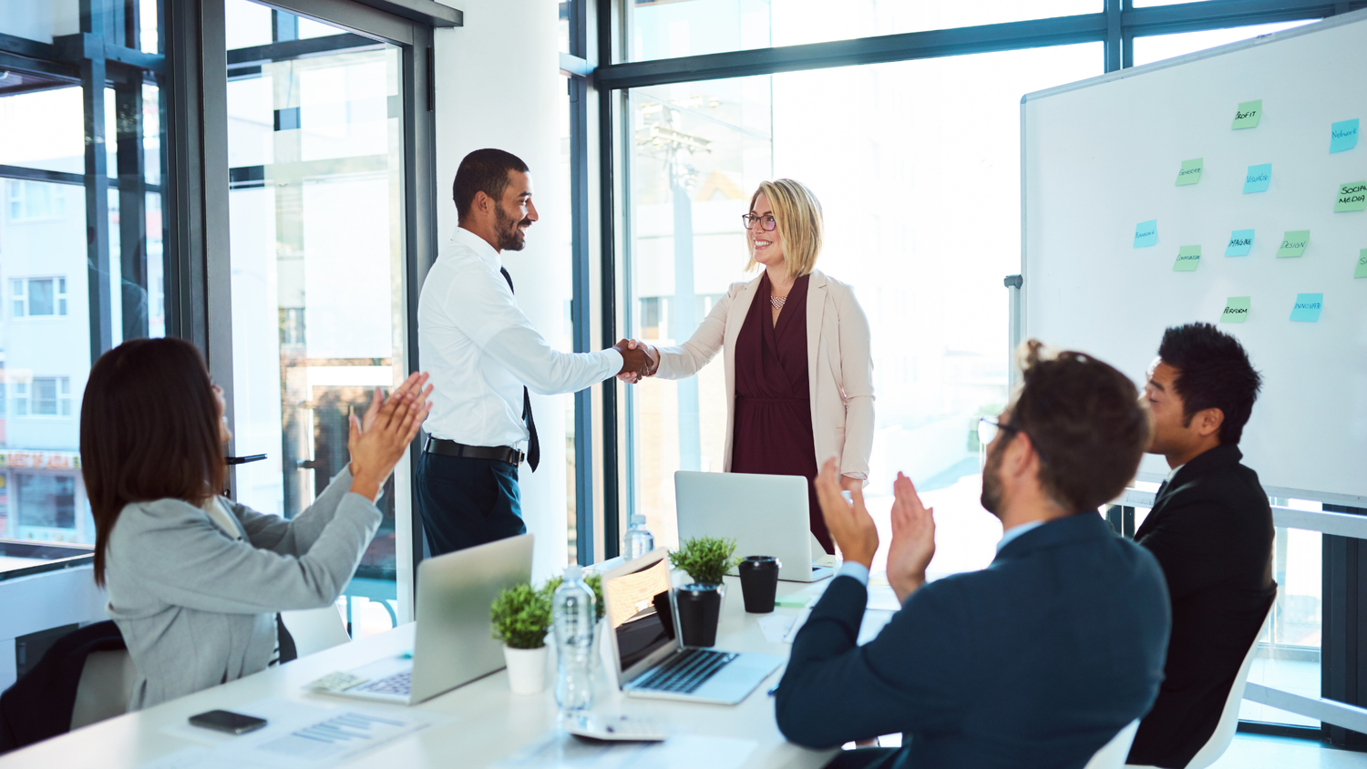 A man and a woman shake hands in a meeting room as colleagues clap, a whiteboard with sticky notes visible.