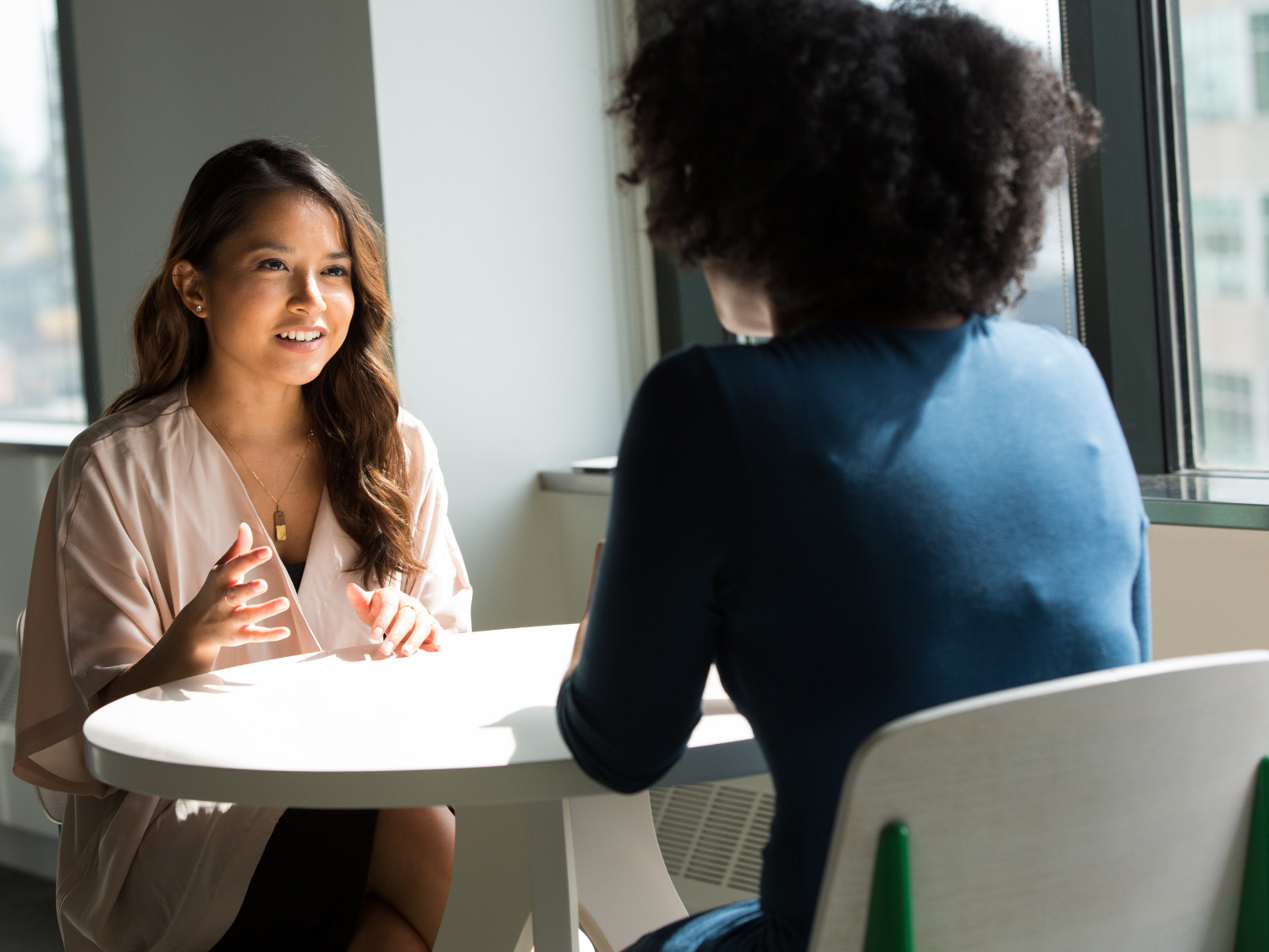 Two women have a discussion across a white table; one facing the camera gestures as she speaks.