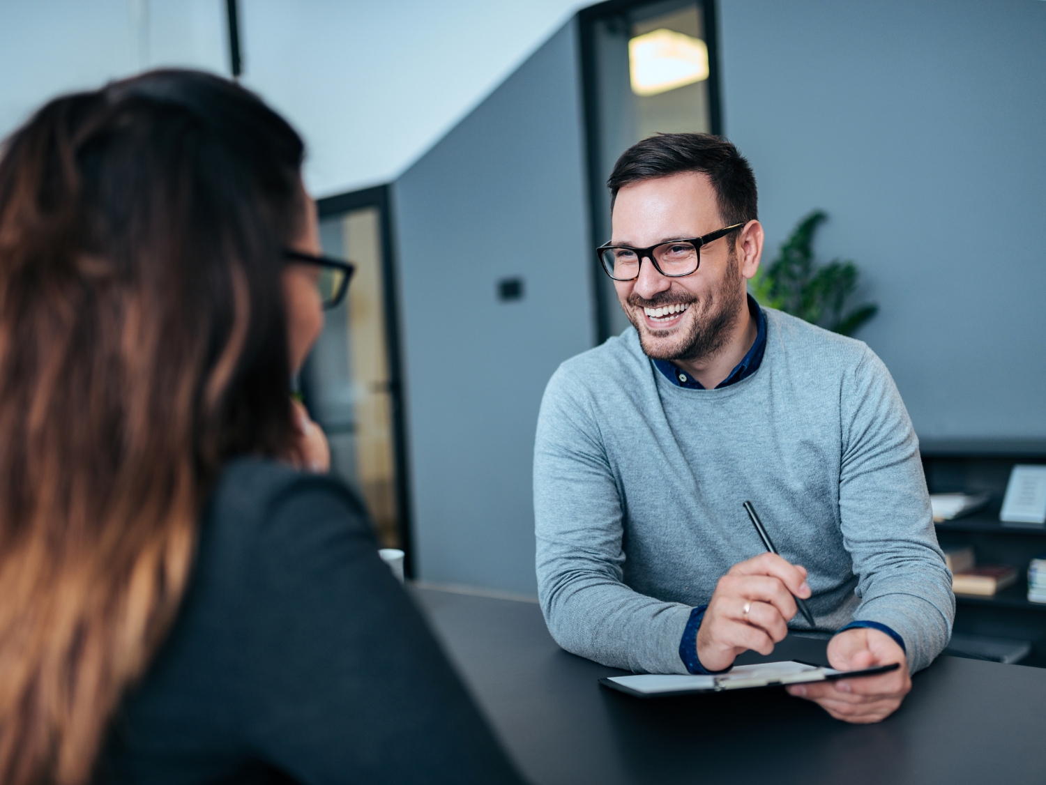 Smiling man in glasses holds a pen over a clipboard while looking at a woman across a table.