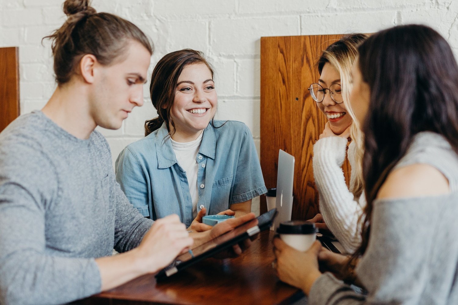 Four young adults gathered around a table interacting with technology; one man holds a tablet while others look on.