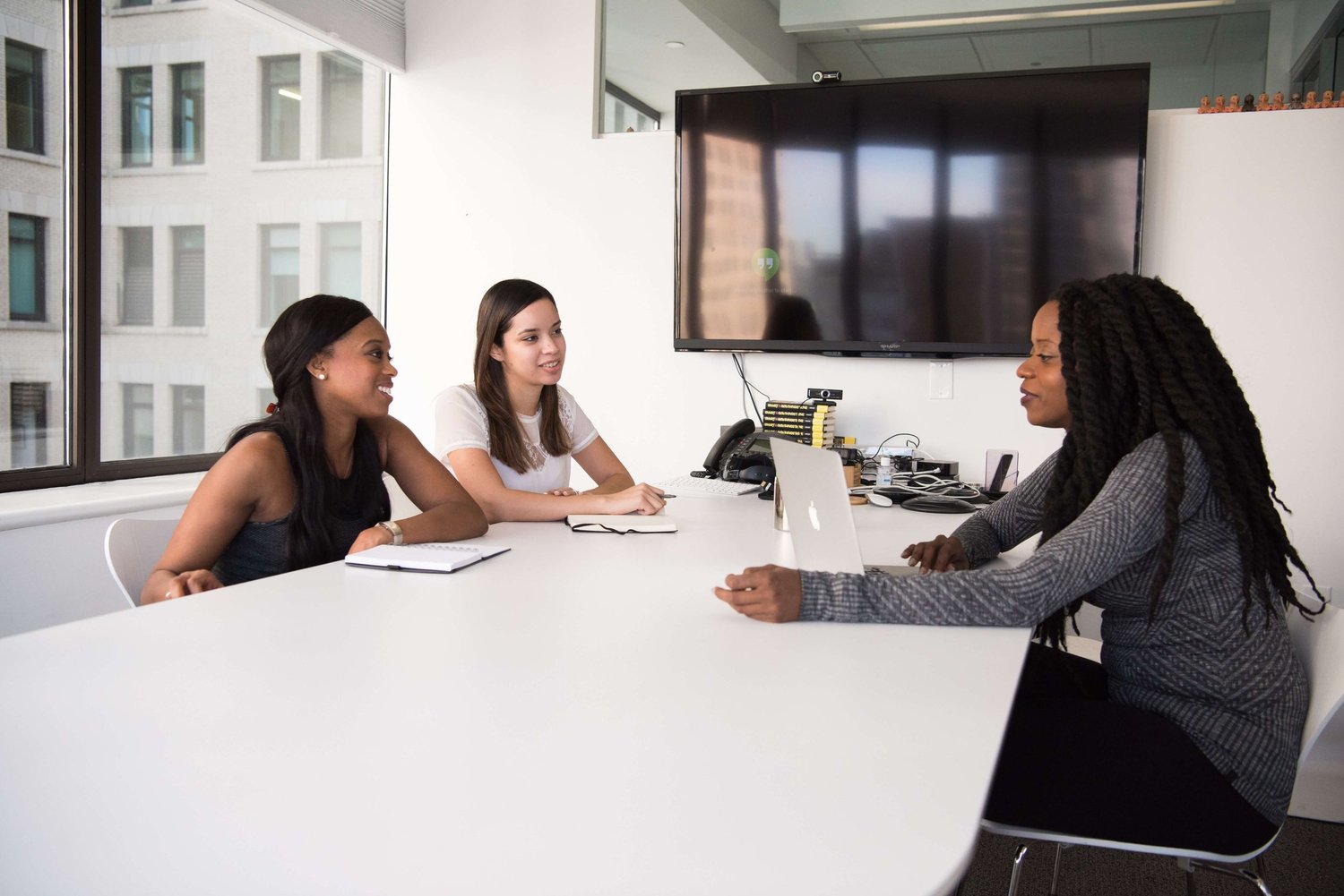 Three women at a conference table in a modern office; the woman on the left smiles toward the center.
