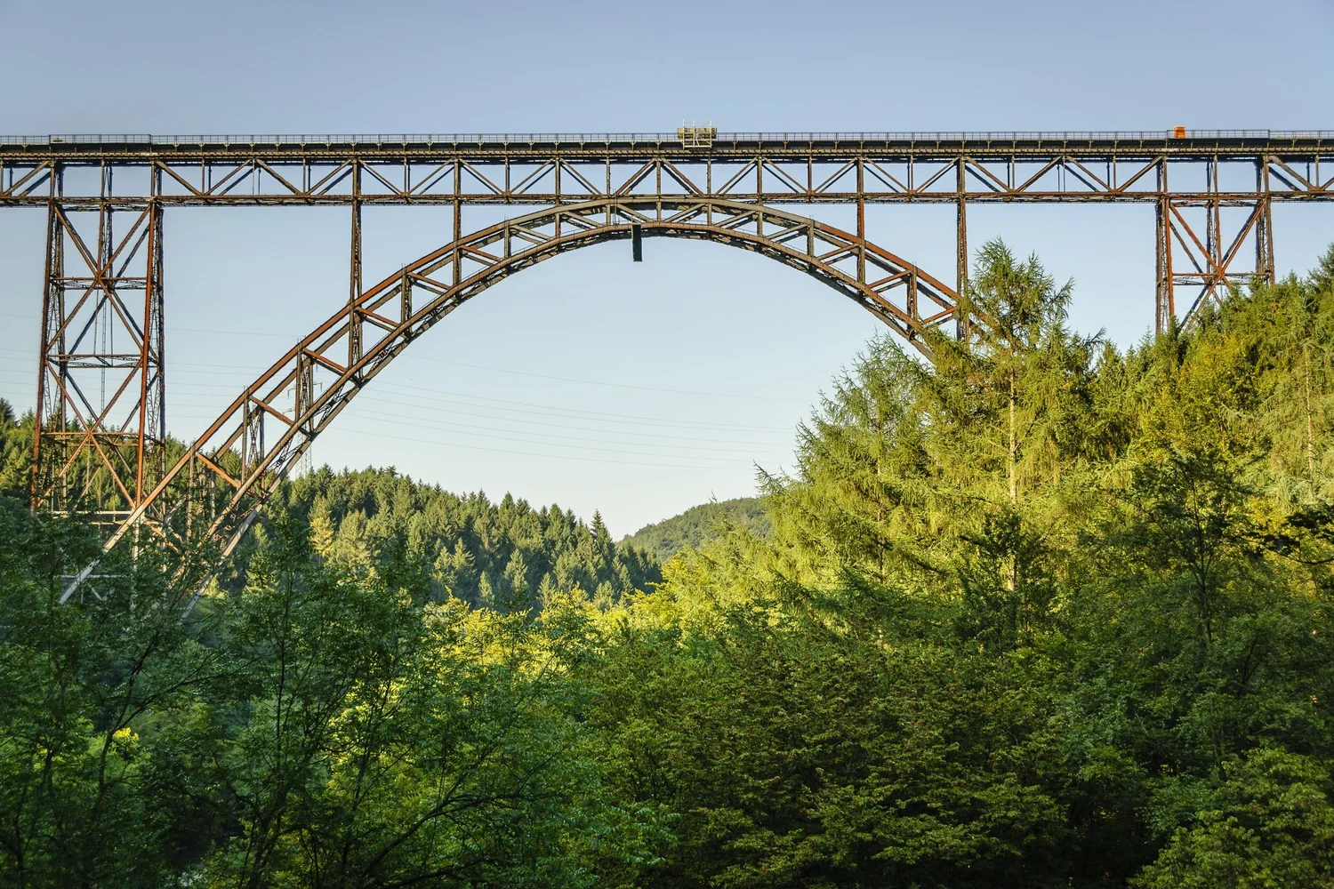 A large, rust-colored metal arch bridge spans a forested valley under a clear blue sky.