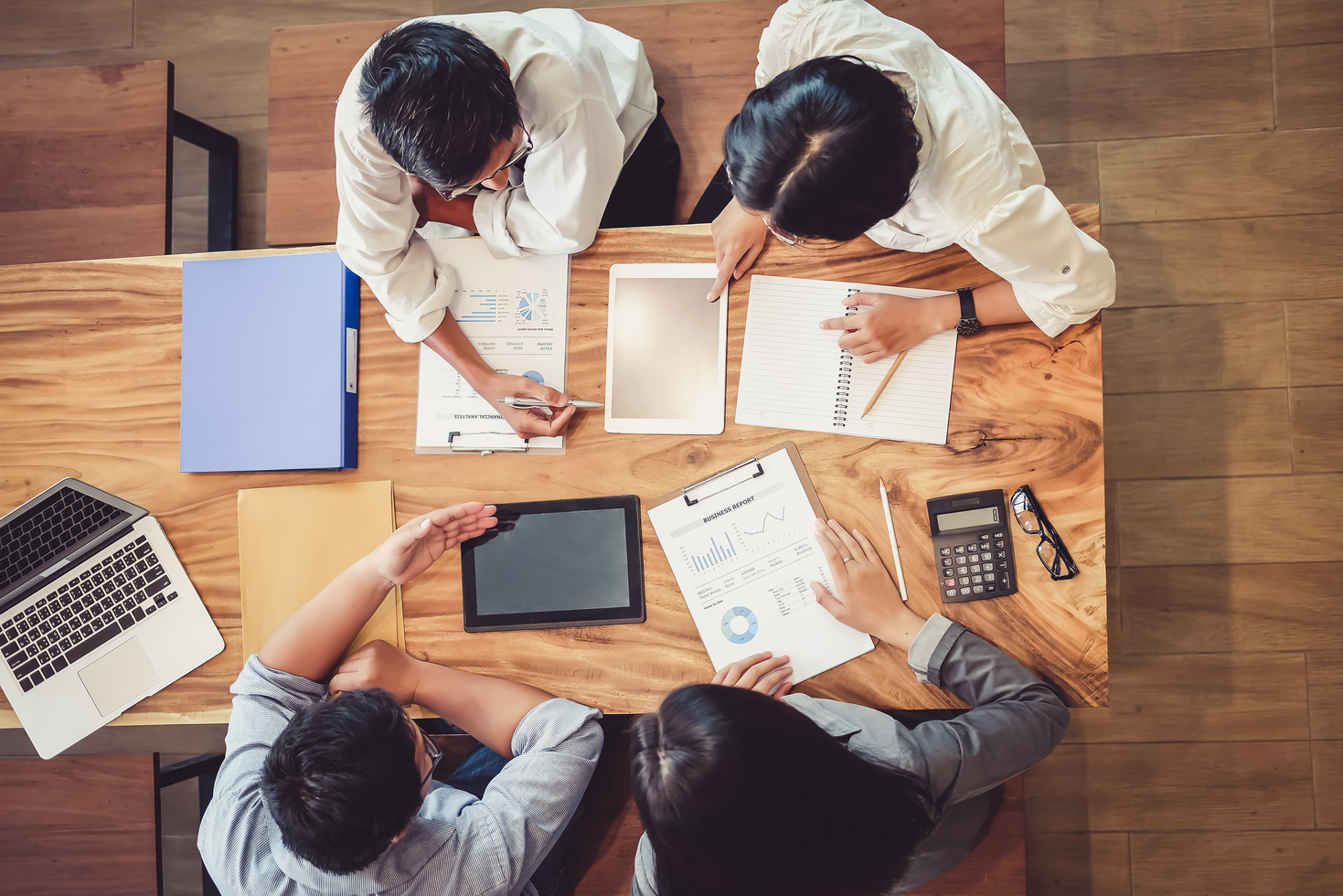 Four colleagues seen from above around a wooden table, reviewing printed charts, a business report, a tablet and a laptop